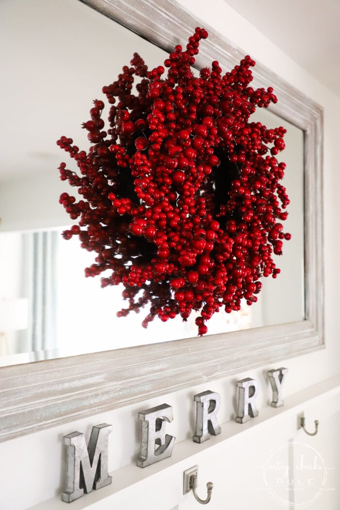 white rustic wall mirror with red berry wreath hung over the top of itunderneath is the letters MERRY in wooden rustic ornaments.